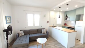Un apartamento moderno con un sofá seccional gris, una mesita redonda, una cocina con refrigerador blanco y un ventanal grande.