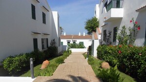 Un sendero conduce a un edificio de paredes blancas y contraventanas verdes, rodeado de jardines cuidados y un cielo despejado.