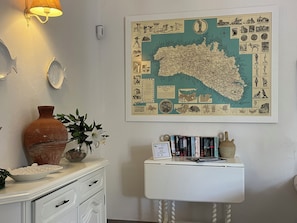 Ett rum med en karta på väggen, en vit byrå, en vas och ett litet bord med böcker.