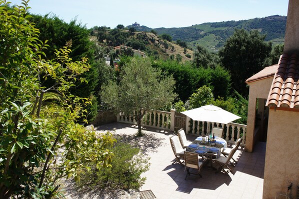 A patio with outdoor furniture and a table, surrounded by greenery and hills in the background.