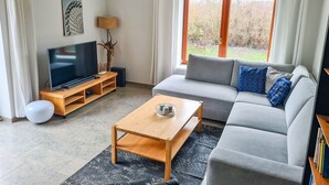 Ein modernes Wohnzimmer mit einem grauen Sofa, einem Holztisch, einem Fernseher auf einem Holzschrank und einem großen Fenster mit Blick auf Bäume.
