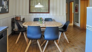 Ein Essbereich mit einem runden Holztisch, sechs Stühlen, einer hängenden Pendelleuchte und einer Küchenzeile mit Kühlschrank und Herd.