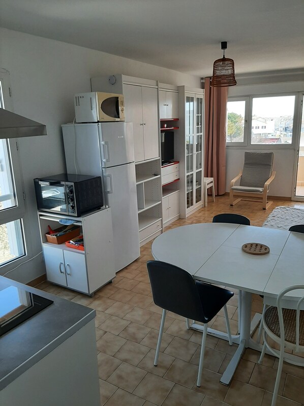 A compact kitchen with white appliances, a dining table with chairs, and a view through a window.
