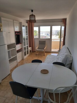 A compact living space with a round dining table, a sofa, a chair, a wardrobe, and a balcony with a view of buildings.