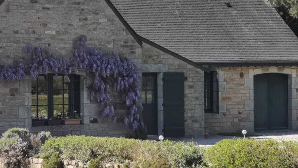 Edifici de pedra amb persianes verdes, un porxo amb plantes en testos i enfilades de flors morades.