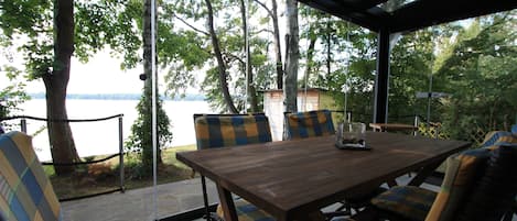 Una mesa de madera con sillas a cuadros y una lámpara de cristal, frente a un ventanal con vistas a un lago.