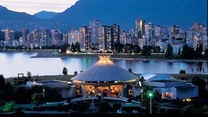Paisagem urbana com um edifício de destaque, apresentando uma grande estrutura em cúpula iluminada em primeiro plano, cercada por um corpo de água e uma paisagem montanhosa ao fundo.
