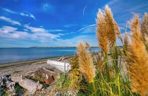 Une plage bordée d’un rivage rocheux, un petit bateau et de hautes herbes au premier plan.