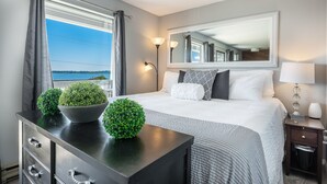 Quarto bem arrumado com uma cama grande, cômoda com plantas decorativas, abajur e vista para o exterior.