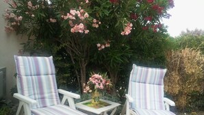 Two white chairs with striped cushions, a small table with a vase of flowers, and a garden with various plants and trees. - COMFORTABLE SEA-FRONT PAVILION (Fleury)