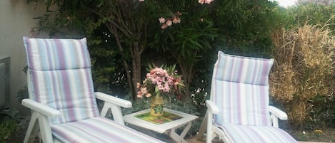 Zwei weiße Stühle mit gestreiften Kissen, ein kleiner Tisch mit einer Blumenvase und ein Garten mit verschiedenen Pflanzen und Bäumen.