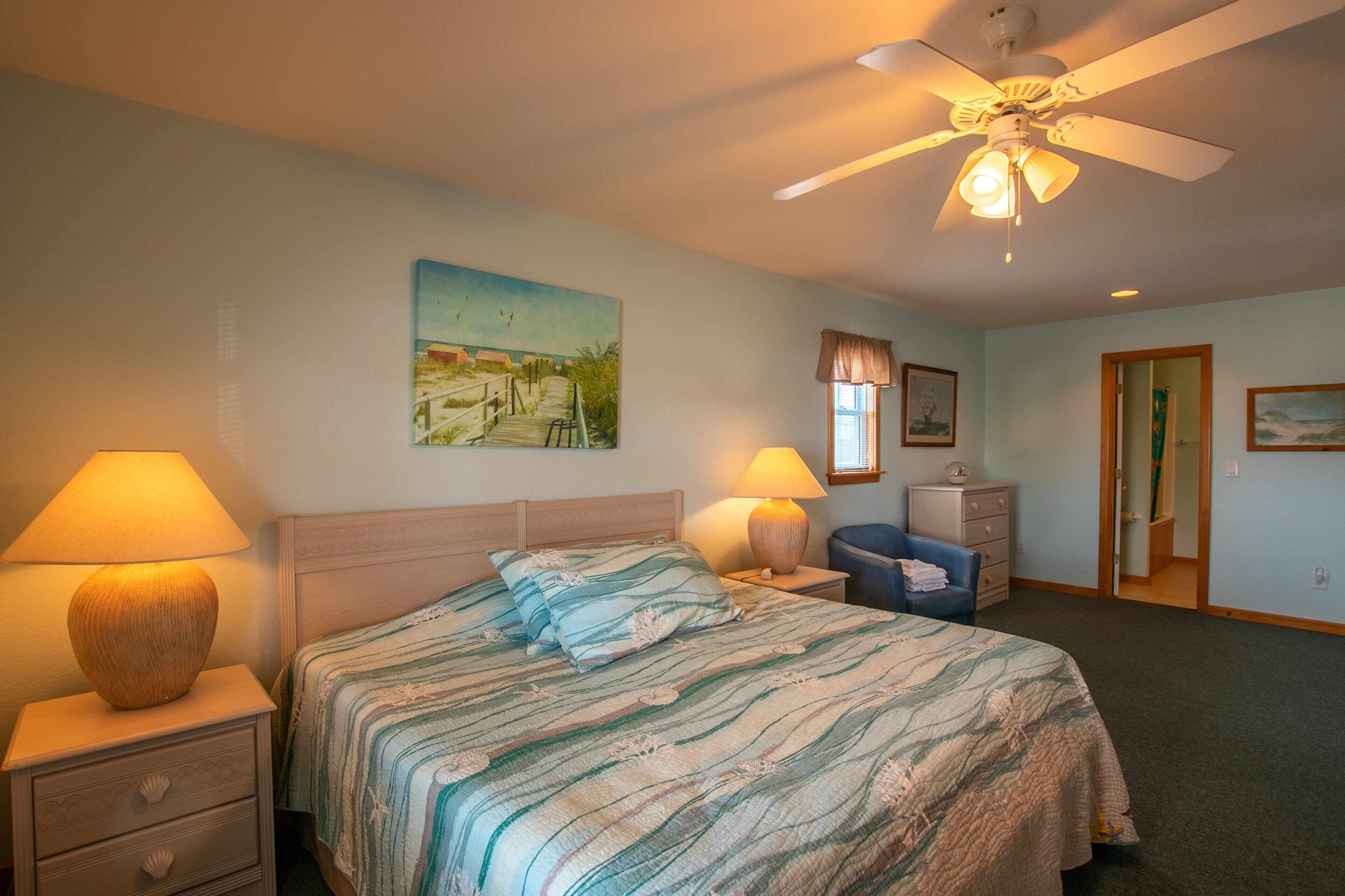 A bedroom with a bed, two bedside tables, a ceiling fan, a window with curtains, a nightstand, and a painting on the wall.