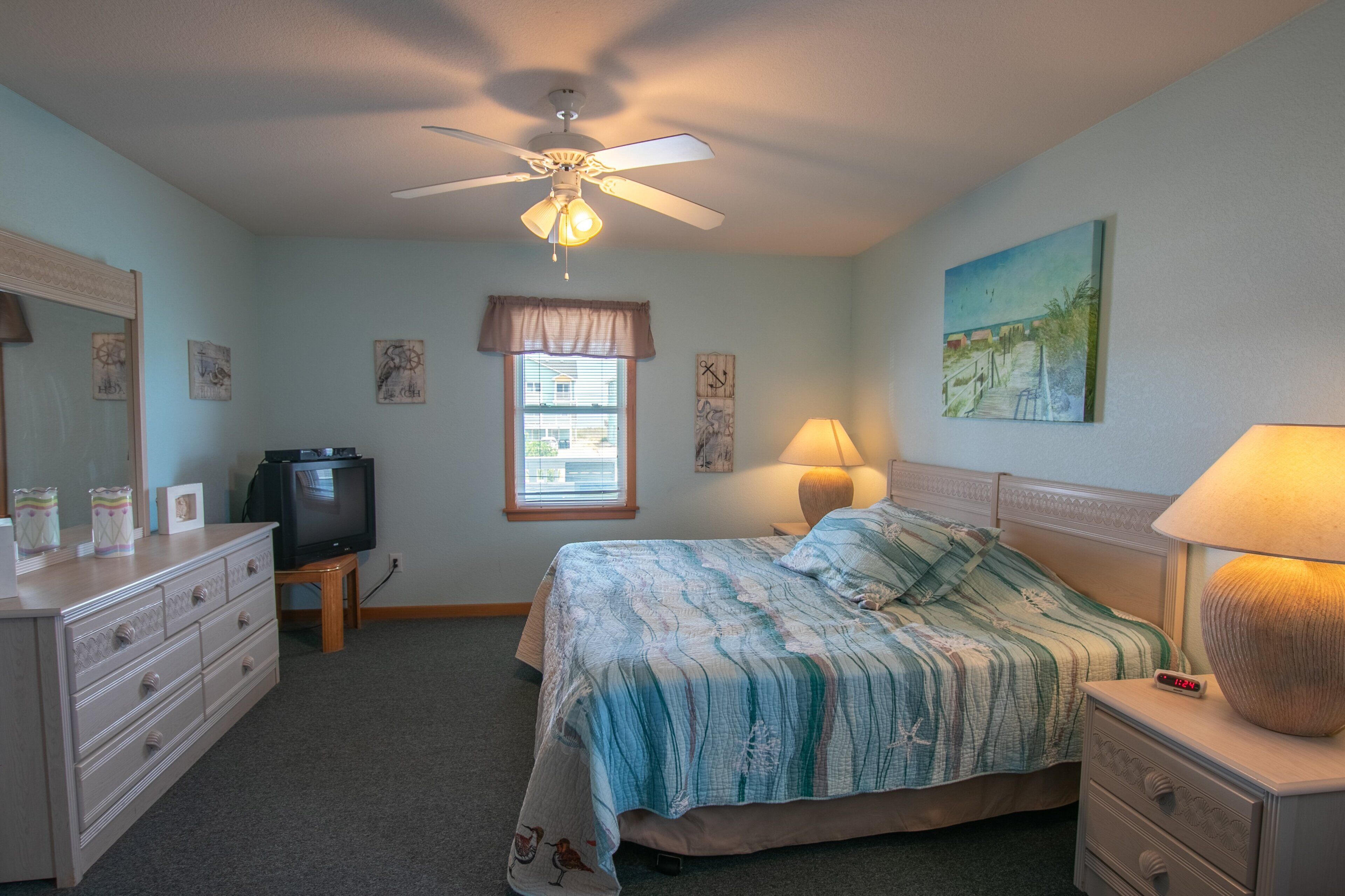 Une chambre à coucher comprenant un lit, une commode, un téléviseur, un ventilateur de plafond, une fenêtre et des œuvres d’art.