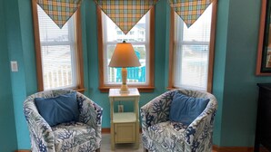 A room with teal walls, two patterned armchairs with blue cushions, a small table with a lamp, and a window with blinds. - 5454- Sea View - Nags Head Semi-Oceanfront | Private Pool (Nags Head)