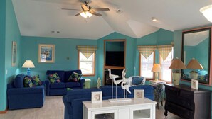 A living room with teal walls, blue sofas, a white cabinet, and a ceiling fan. - 5454- Sea View - Nags Head Semi-Oceanfront | Private Pool (Nags Head)