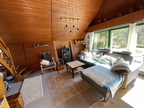 A cozy cabin interior with a wooden ceiling, a sofa, a chair, a small table, and a window offering a view of the outdoors.