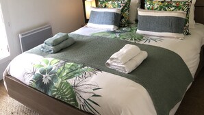 Una cama bien hecha con estampado de hojas tropicales, almohadas verdes y blancas, y un cuadro en la pared.