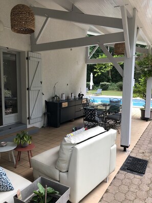 A covered patio area with a white sofa, a small table with a plant, a black cabinet, and a wicker hanging light fixture.
