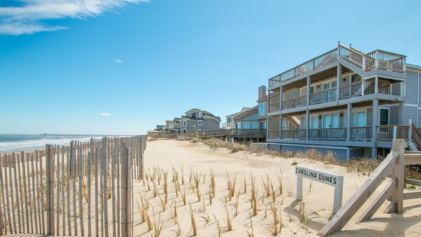 A beachfront property with a wooden fence, a sign reading "CAROLINA DUNES," and a multi-story building with balconies. - 2875 - MOJO'S, Duck Beachfront Home w/ Pool Table & Views (Duck)