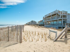 A beachfront property with a wooden fence, a sign reading "CAROLINA DUNES," and a multi-story building with balconies. - 2875 - MOJO'S, Duck Beachfront Home w/ Pool Table & Views (Duck)