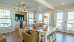 A spacious living room with a beige sofa, a coffee table, and a TV area with cabinets. There are ceiling fans, large windows with a view of the beach, and a door leading to the outside. - 2875 - MOJO'S, Duck Beachfront Home w/ Pool Table & Views (Duck)