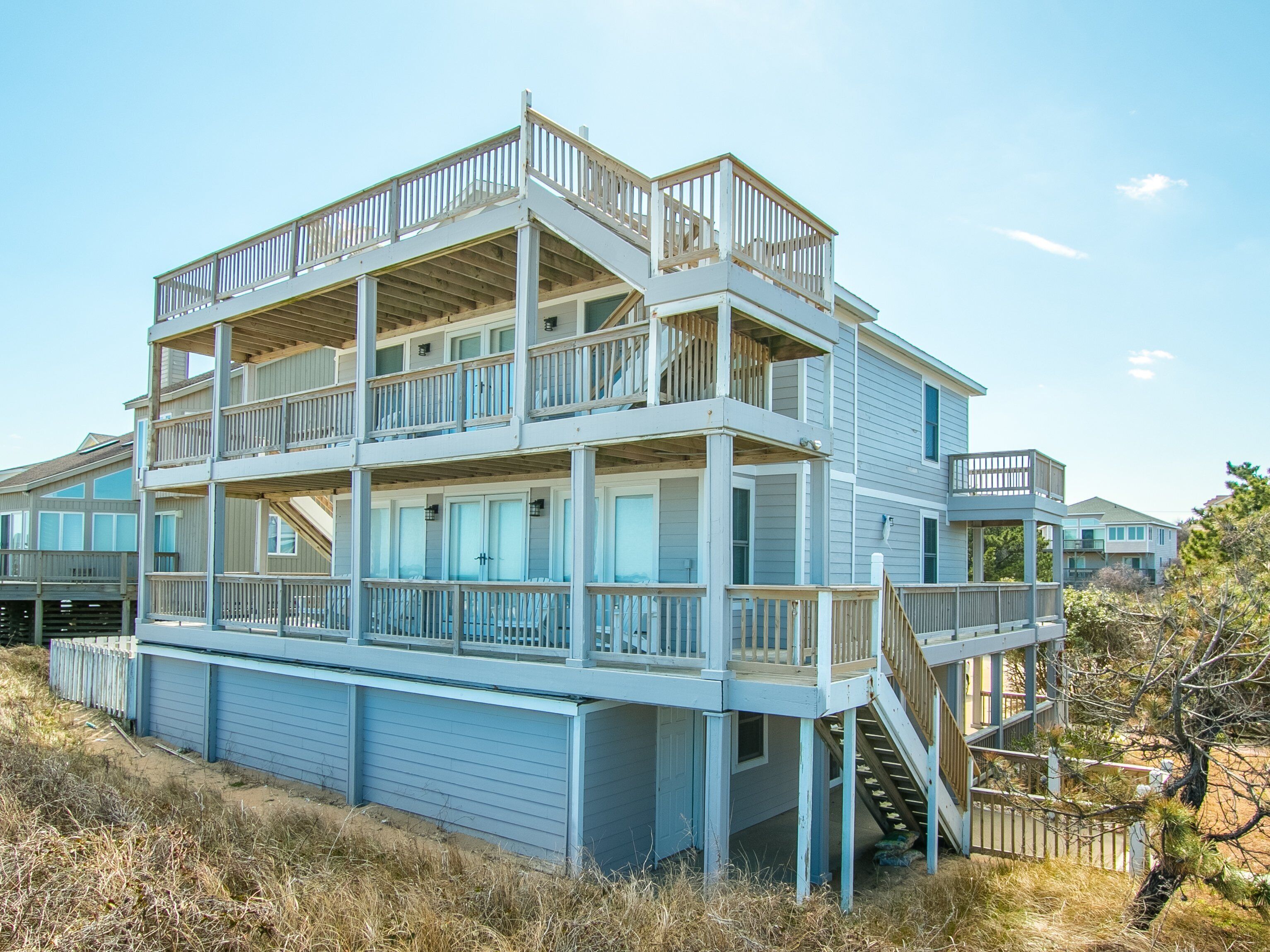 Une maison à plusieurs étages située en bord de mer, dotée d’une terrasse et d’un balcon en bois, et comprenant un escalier menant aux niveaux supérieurs.