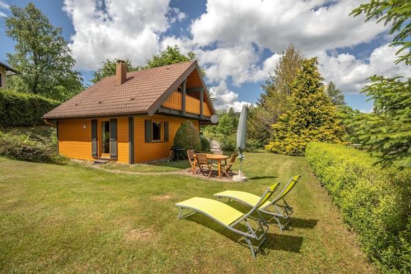 A yellow wooden house with a red roof, a balcony, and a lawn with two yellow lounge chairs.