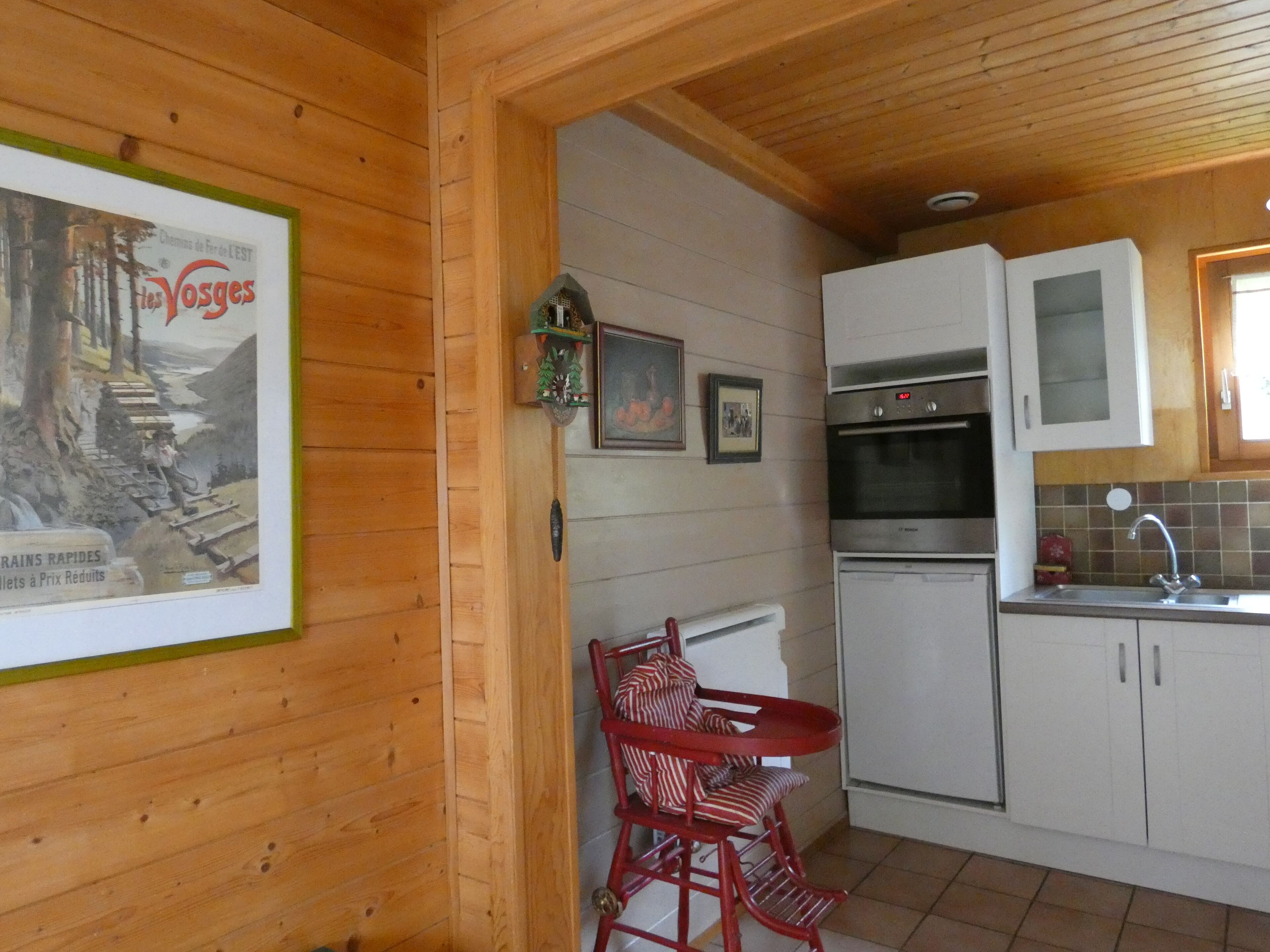 A wooden interior with a kitchen area including a stove, oven, and sink. There is a framed picture on the wall and a small table with red chairs.