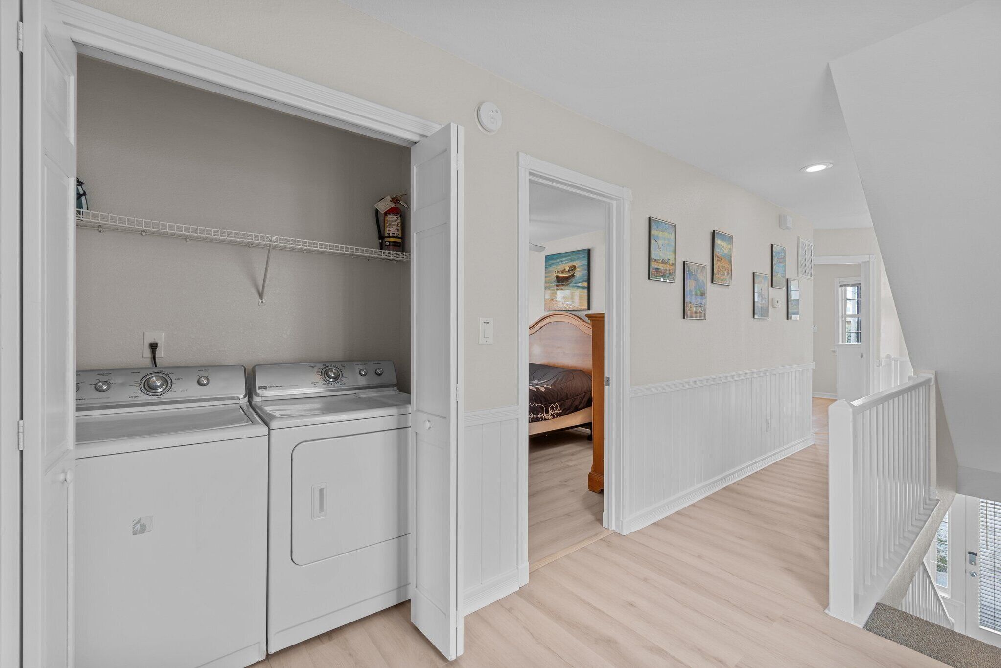 A laundry room with stacked washer and dryer units, a shelf with toiletries, and a hallway leading to a bedroom with a bed and artwork on the wall.