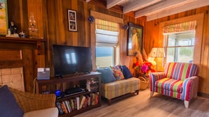 A cozy living room with wooden walls, a television, a bookshelf, a striped armchair, and a window with a view. - 5452- Sea Captain's Cottage - Historic Oceanfront Cottage | Nags Head (Nags Head)