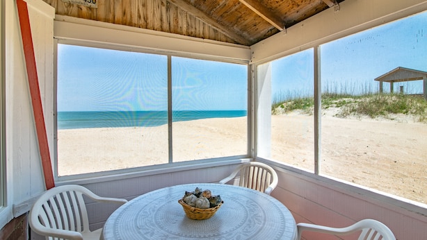 A covered outdoor seating area with a view of the beach, a table with a basket of items, and a wooden structure in the background. - 5452- Sea Captain's Cottage - Historic Oceanfront Cottage | Nags Head (Nags Head)