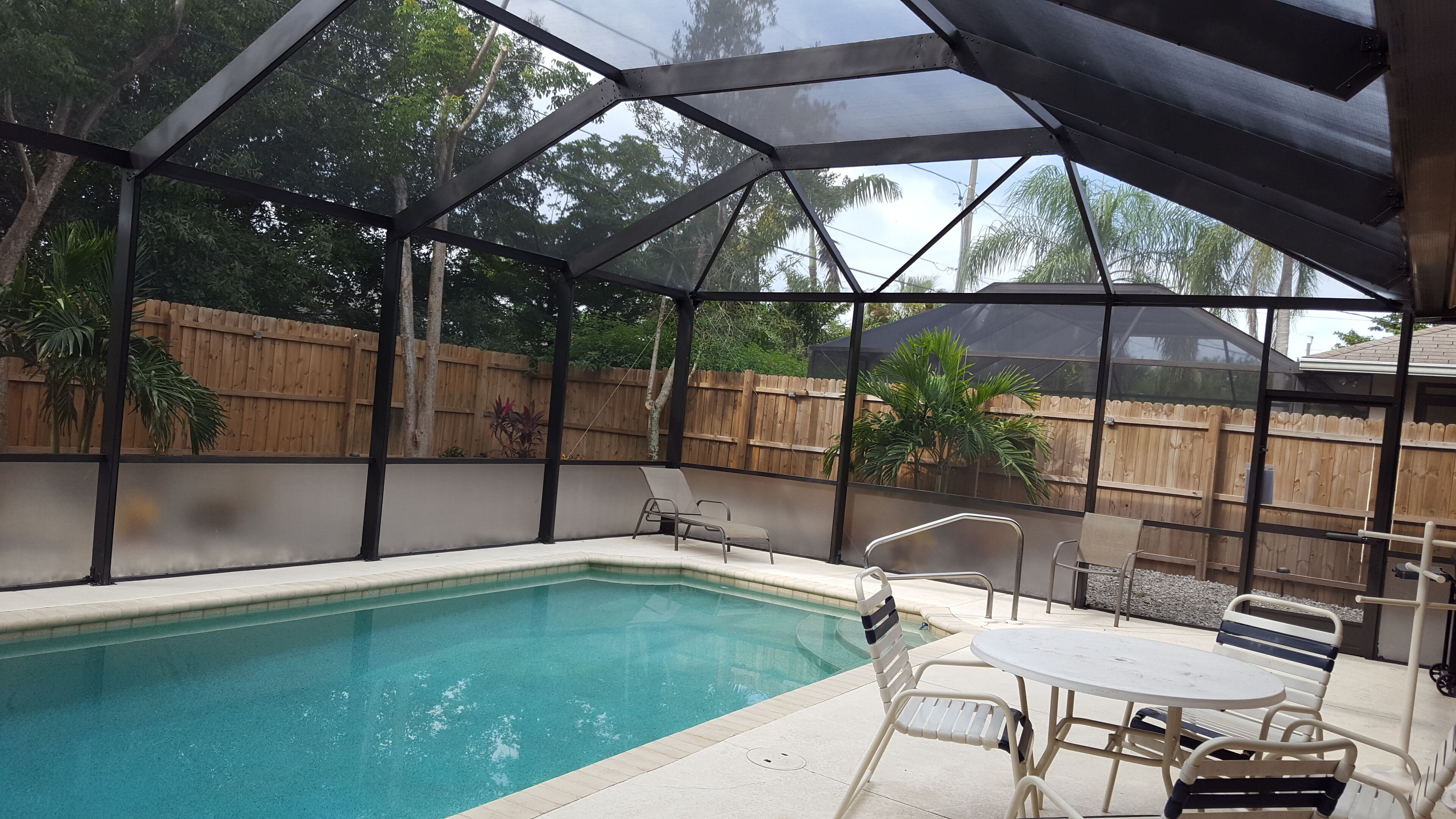 A covered outdoor swimming pool with a wooden fence, lounge chairs, and a table.