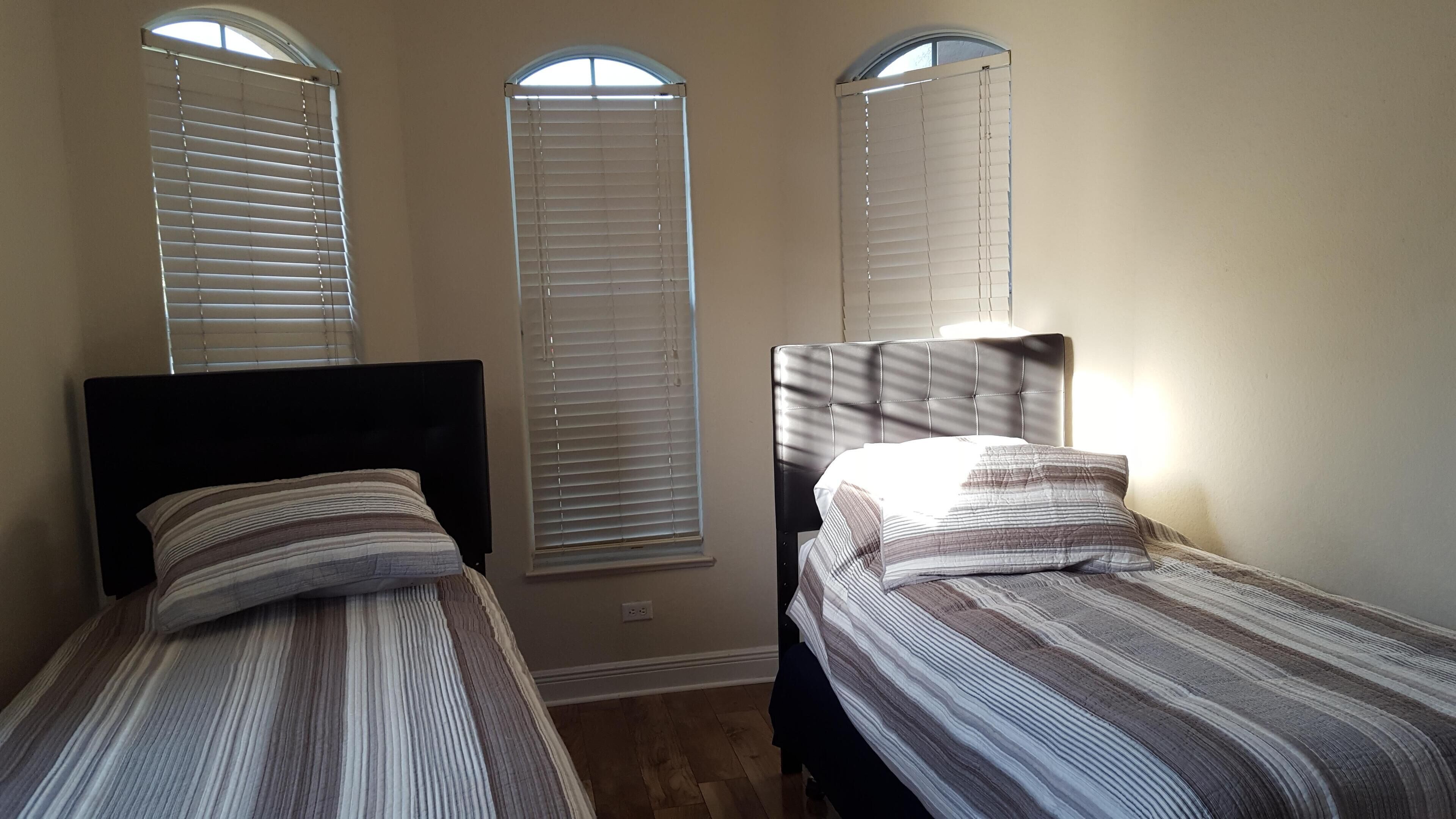 A room with two beds, each with a headboard and a striped bedspread. There are three windows with blinds, and the room has a wooden floor.