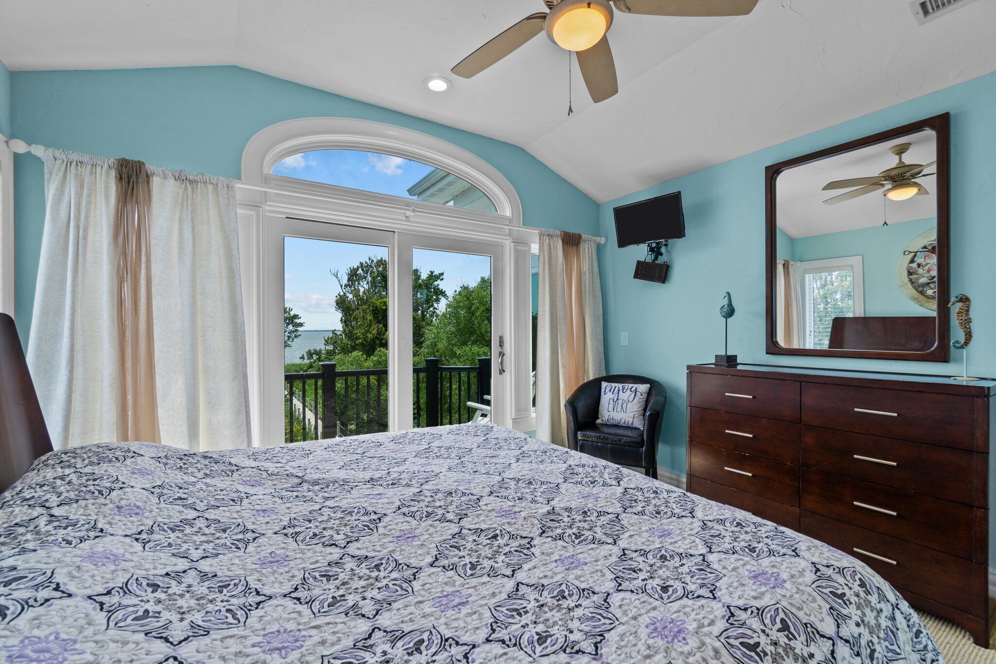 A bedroom with a large bed, a dresser, a chair, a ceiling fan, and a balcony with a view of trees.