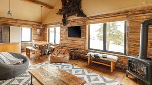 Interior de una cabaña de troncos con un gran cráneo de oso en el techo, muebles de madera, una chimenea y ventanas amplias.