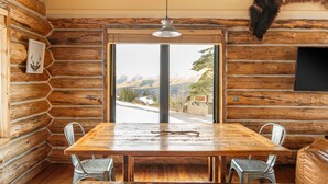 Un interior rústico de madera con una gran mesa de comedor, sillas de metal, una cabeza de animal montada y vistas a un paisaje nevado a través de un ventanal.