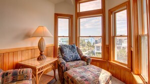 A cozy corner with a cushioned chair, a small table with a lamp, and large windows offering a view of neighboring houses. - 1600 - Bases Loaded - Spacious Buck Island Retreat in Corolla (Corolla)