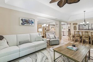 A spacious living room with a sofa, armchair, coffee table, and dining area with chairs and a chandelier.