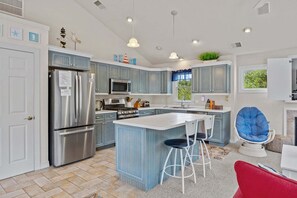 A modern kitchen with blue cabinets, stainless steel appliances, a central island with stools, and a hanging chair. - 1820 - The Thirsty Duffer, Currituck Club Retreat | Pool, Hot Tub & Golf (Corolla)