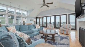 A spacious living room with a large sofa, a round coffee table, and striped armchairs. There is a ceiling fan, large windows offering a view of the outdoors, and a flat-screen TV mounted on the wall. - 5464- EY BELIEVE - Oceanfront Nags Head | 8BR Luxury Retreat (Nags Head)