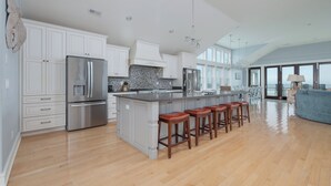 A spacious kitchen with white cabinetry, stainless steel appliances, and a large island with bar stools. - 5464- EY BELIEVE - Oceanfront Nags Head | 8BR Luxury Retreat (Nags Head)
