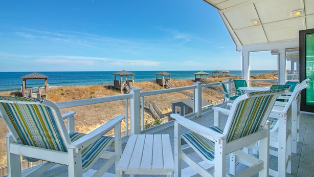 A beachside deck with white chairs and a table, overlooking the ocean and beach huts. - 5464- EY BELIEVE - Oceanfront Nags Head | 8BR Luxury Retreat (Nags Head)