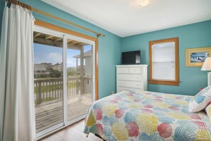 Une chambre avec une grande porte-fenêtre coulissante donnant sur un balcon, une commode blanche, un téléviseur posé dessus, et un lit recouvert d’une couverture aux couleurs vives.