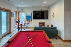 A living room with a pool table, a sofa, a television, and a view of the outdoors through large windows. - 5405- Adagio - Oceanfront Nags Head | Pool, Hot Tub & Elevator (Nags Head)