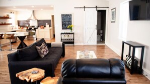 Una sala de estar moderna con un sofá de cuero negro, una mesa de centro de madera y un sillón de cuero negro. Se ve una cocina con gabinetes blancos y una zona de comedor con taburetes de bar al fondo.