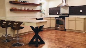 Una cocina moderna con piso de madera, una isla central, taburetes de bar, horno empotrado y extractor de acero inoxidable.