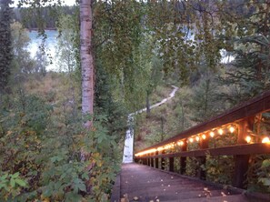 A wooden bridge with lights, surrounded by trees and a path leading to a building. - Kenai Riverfront Vacation Cottage  (Soldotna)