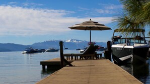 A wooden dock with lounge chairs and an umbrella, boats docked in the background, and mountains in the distance.