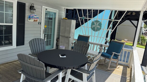 En overdækket terrasse med et rundt bord og fire stole, en dør med et skilt, og en blå stol med pude.