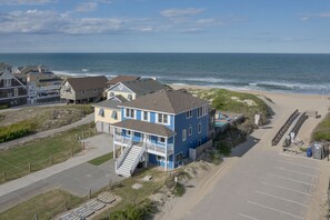 A coastal building with a staircase leading to a deck, adjacent to a beach with a parking area. - 5514- Sisterly Love - Luxury Oceanfront | Pool, Hot Tub & Views (Nags Head)